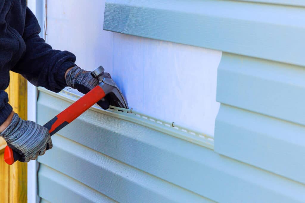 - A professional contractor using a hammer to securely install light blue new siding panels.